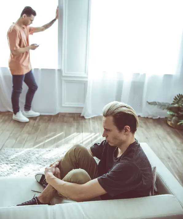 Toned portrait of unhappy guy with dyed hair sitting on couch while his boyfriend standing near window and using smartphone